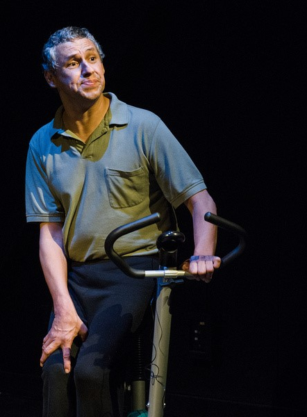Sam Valdez, a light-skinned Mexican-American man with salt and pepper hair sits on an exercise bike, lit dramatically by stage lights.